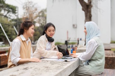 Aralarında tesettürde bir kişinin de bulunduğu üç kadından oluşan çeşitli bir grup, açık hava projelerinde işbirliği yapıyor, takım çalışmasını teşvik ediyor ve akademik bir ortamda fikirleri paylaşıyor..