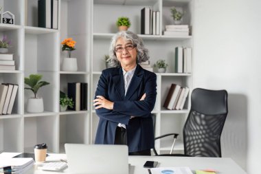Gri saçlı, kendinden emin bir üst düzey işletme yöneticisi modern bir ofiste kollarını kavuşturarak organize ofis malzemeleri arasında profesyonellik ve liderlik yayıyor..