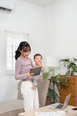 Becerikli bir anne bebeğiyle kollarında duruyor, bir tabletle uğraşıyor, teknolojinin modern ebeveynlik ve iş hayatı dengesine entegrasyonunu anlatıyor..
