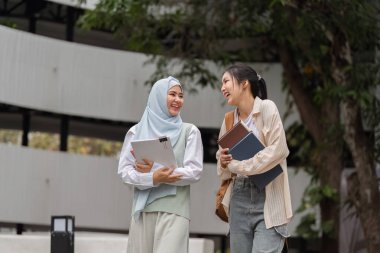 Kampüste birlikte yürüyen, gülümseyen ve gülüşen iki farklı genç kadın eğitim materyallerini tutarken ve akademik deneyimlerini tartışırken.