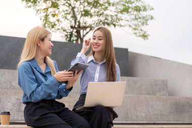 Açık hava ortamında tablet ve laptop kullanarak fikir alışverişinde bulunan iki farklı kadın, takım çalışmasını ve yeniliği vurguladı.