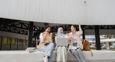 Heyecanlı çeşitli bayan öğrenciler akademik başarılarını kutluyorlar, bir dizüstü bilgisayar kullanırken sevinçle ellerini kaldırıyorlar ve üniversite ortamında kitaplarla çevrelenmişler..