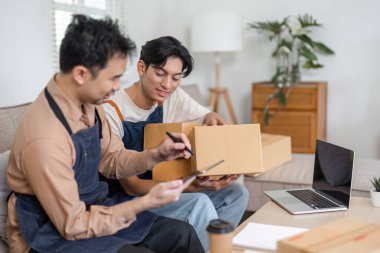 İki adam internetteki satışları için öngörülerini paylaşıyor ve kutuları paketliyor. Oturma odalarında düzenli bir çalışma alanıyla çevrili..