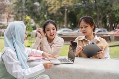 Çeşitli üniversite öğrencilerinden oluşan bir grup, park benzeri bir ortamda dizüstü bilgisayar ve tablet kullanırken fikir alışverişinde bulunuyorlar..