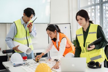 Çeşitli bir inşaat ekibi, parlak, modern bir ofis alanının araç ve planlarını kullanarak bina tasarımlarını aktif olarak planlar ve ölçer..