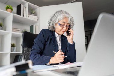 Yaşlı bir iş kadını sorumluluklarını yerine getiriyor, telefonda konuşuyor ve şık bir ofis ortamında notlar alıyor..