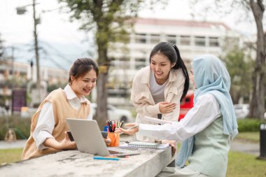 Açık havada dizüstü bilgisayarın etrafında toplanan, fikirleri paylaşan ve üniversite ortamında işbirliği yapan çok kültürlü bir kadın grubu..