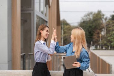 İki farklı kadın başarılarını bir beşlikle kutlarken açık havada bir tablet tutuyor, neşe ve takım çalışması sergiliyorlar..