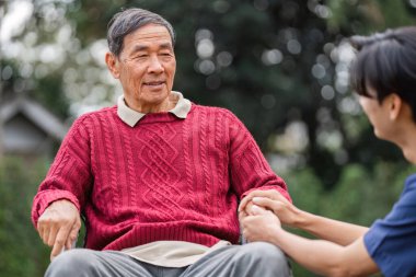 Rahat bir süveter giyen yaşlı bir adam bakıcısıyla sıcak bir sohbete dalmış, huzurlu bir bahçe ortamında güzel bir günün tadını çıkarıyor..