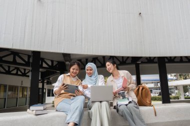 Bir grup çok kültürlü kız öğrenci, kampüs ortamında kitaplarla çevrili bir tablet ve dizüstü bilgisayar kullanarak fikir paylaşıp işbirliği yapıyorlar..
