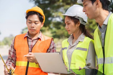 Aralarında bir mühendis ve bir denetçinin de yer aldığı üç inşaat işçisi, planları analiz ediyor ve bölgede ölçümleri tartışırken, kasklar ve yeleklerle güvenliği sağlıyorlar..
