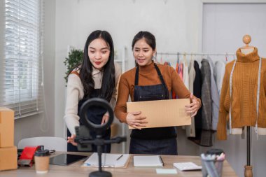 Çevrimiçi satışlara aktif olarak hazırlanan iki kadın, giysi ürünlerini düzenliyor ve canlı giysilerle dolu şık bir çalışma alanında ürünleri paketliyorlar..