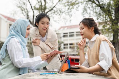 Bir kampüs ortamında dizüstü bilgisayar ve çeşitli kırtasiye ürünleri kullanırken üç farklı kız öğrenci notlarını paylaşıyor ve dışarıda fikirleri tartışıyorlar..