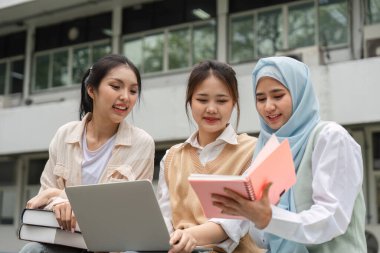 Bir grup farklı öğrenci bir üniversite kampüsünde otururken bir dizüstü bilgisayar ve defterle birlikte gülümsüyor ve ders çalışıyor..