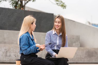Modern bir açık hava çalışma sahasında laptopla sohbet eden ve işbirliği yapan iki farklı kadın, takım çalışmasını ve yaratıcılığı teşvik ediyor..