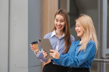İki farklı kadın bir tablet üzerinde gülümsüyor ve işbirliği yapıyor. Bu arada biri kahve tutuyor, modern ofis ortamında takım çalışması sergiliyor..