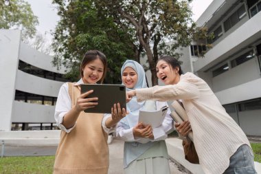 Üç farklı çok kültürlü kadın kampüsteki bir tablette heyecanlı bir şekilde bilgi izliyorlar, destekleyici bir ortamda kahkaha ve içgörü paylaşıyorlar..