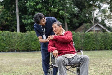 Bir sağlık çalışanı, tekerlekli sandalyedeki yaşlı bir adamla konuşmak için öne eğiliyor. İkisi de sakin bir bahçe ortamında gerçek bir bağ kuruyor..