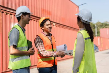 Üç inşaat işçisi el sıkışırken, bir inşaat sahasında konteynırların yakınındaki proje planlarını tartışırken ortaklığı sağlamlaştırdılar..