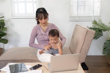 Şefkatli bir anne iş yerinde oturur, bebeğiyle neşeyle uğraşırken bilgisayarına odaklanır, rahat bir evde ilham verici bir iş ve ebeveynlik dengesi yaratır..