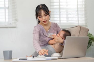 Çalışkan bir anne bebeğini biberonla besler ve görevlerini parlak ve modern bir ofis ortamında dizüstü bilgisayarla yönetir..