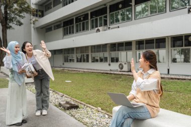 Çeşitli öğrencilerden oluşan bir grup, üniversite kampüsünde dizüstü bilgisayarla oturan bir arkadaşına el sallıyor ve selam veriyor..