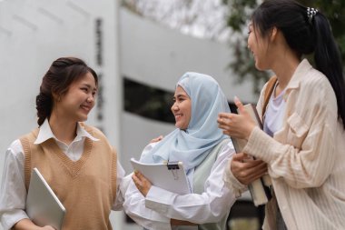 Üç farklı genç kadın birlikte dışarıda vakit geçiriyorlar, gülümsüyorlar ve öğrenim materyallerini paylaşıyorlar. Arkadaşlıklarını ve akademik yolculuklarını kutluyorlar..