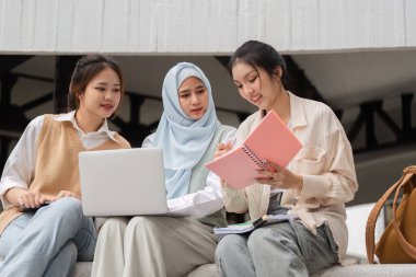 Bir dizüstü bilgisayar kullanarak ve akademik çalışmalarını yaparken notlarını paylaşarak işbirliği içinde dışarıda okuyan üç farklı kız öğrenci..