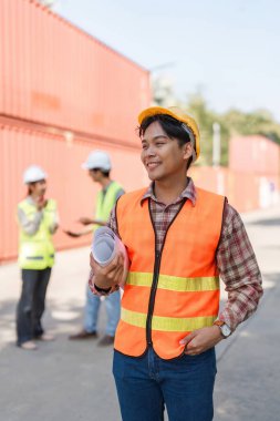 Turuncu güvenlik yeleği ve kasketi olan kendine güvenen bir inşaat mühendisi, şantiyede iş arkadaşlarının tartışmalarını izleyen planlarını gururla elinde tutuyor.