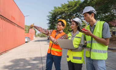 Bir mühendis, bir proje yöneticisi ve bir işçi de dahil olmak üzere çeşitli bir inşaat ekibi, yoğun bir inşaat sahasında dizüstü bilgisayar ve planlar kullanarak proje planları hazırlıyor..
