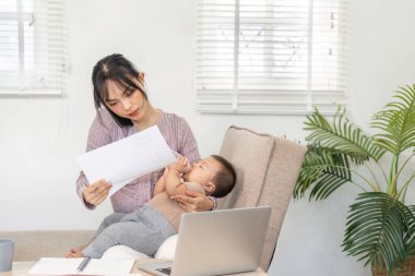Kendini işine adamış bekar bir anne, bebeğini kucağında tutarken iş ve ebeveynliğin rahat bir ev ortamında dengelenmesinin zorluklarını gösteren çalışma belgelerini inceliyor..