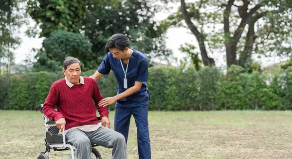 Sakin bir açık hava ortamında, hemşire yardımcısı tekerlekli sandalyedeki yaşlı bir adamı destekliyor ve bakım ve arkadaşlığın önemini vurguluyor..