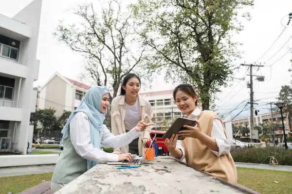 Üç çok kültürlü kız öğrenci açık havada bir projede işbirliği yapıyor, canlı bir kampüs ortamında tablet ve laptop kullanarak fikirleri paylaşıyorlar..