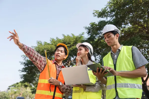 Aralarında bir mühendis ve bir denetçinin de bulunduğu çeşitli inşaat profesyonelleri ekibi açık havada proje detaylarını aktif olarak tartışıyor, dizüstü bilgisayar kullanıyor ve planlara işaret ediyor..