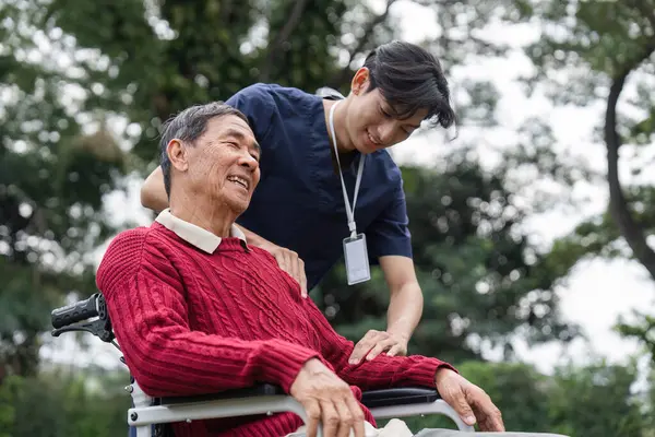 Bir sağlık çalışanı, tekerlekli sandalyedeki gülümseyen yaşlı bir adama nazik bir destek sunarak doğanın güzelliği arasında güven veren bir ilişki sergiliyor..