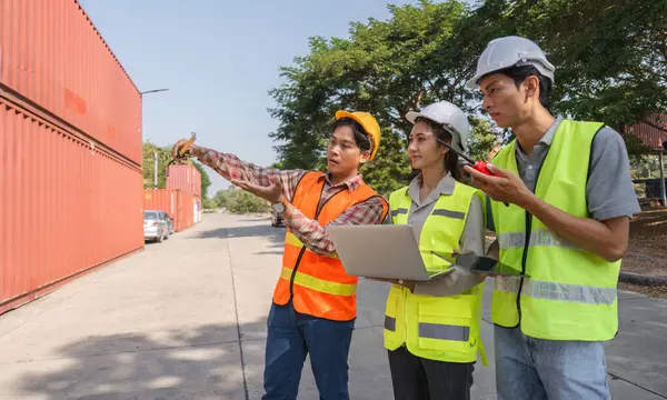 Bir mühendis, bir proje yöneticisi ve bir işçi de dahil olmak üzere çeşitli bir inşaat ekibi, yoğun bir inşaat sahasında dizüstü bilgisayar ve planlar kullanarak proje planları hazırlıyor..
