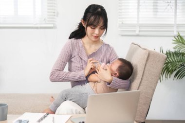Sadık bir anne bebeğine bir şişeyle bakarken ev ofisinin sıcak ve davetkar ortamında laptopunda işini dengeliyor..