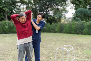 Kendini adamış bir sağlık bakıcısı, huzur dolu bir parkta yürürken yaşlı bir adama süveter giydirerek rehabilitasyona ve desteğe odaklanmasına yardımcı olur..