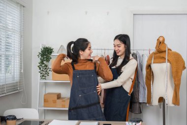 Modern bir stüdyoda, kumaşlar ve aletlerle çevrili iki kadın moda tasarımcısı eğlenceli bir şekilde kıyafet tasarımı üzerinde çalışıyor..