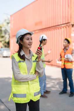 Sert bir şapka ve yansıtıcı güvenlik yeleği giyen kendine güvenen bir kadın inşaat yöneticisi, bir telsiz üzerinden iletişim kuruyor, iş arkadaşlarıyla bir nakliye konteynır deposunda tartışıyorlar..