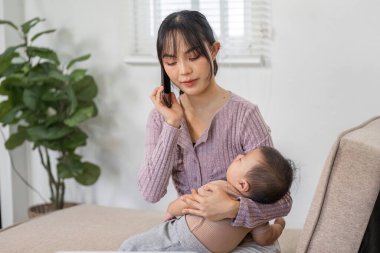 Sorumluluklarını dengeleyen kendini adamış bir anne bebeğini sıcak bir oturma odasında sevgiyle kucaklarken telefonda konuşuyor..
