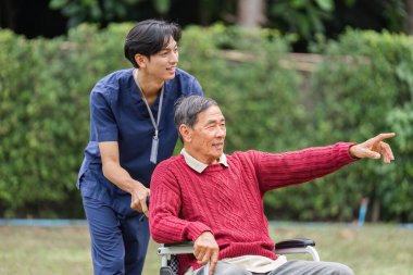 Kırmızı kazaklı yaşlı bir adam, bakıcısı ona tekerlekli sandalyesinde yardım ederken, sevinçle ilgi çekici bir şeyi işaret ediyor.