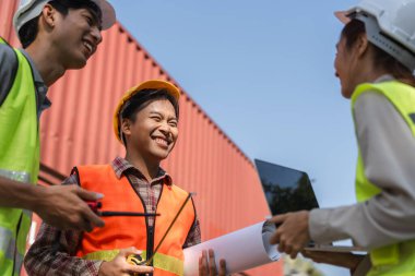 Mühendisler de dahil olmak üzere neşeli bir inşaat ekibi, proje planlarını incelerken ve nakliye konteynırlarının yakınlarındaki ölçümleri tartışırken bir anlık kahkaha paylaşıyor..