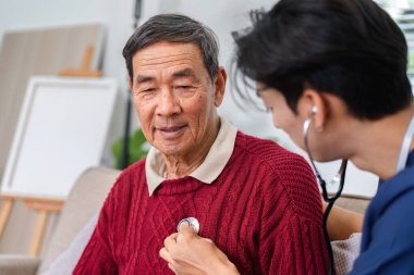 Kendini adamış bir hemşire, oturma odasında steteskopla yaşlı bir adamın kalbini dikkatlice kontrol ederek sağlığını ve sağlığını sağlama alıyor..