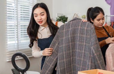 İki kadın şık bir ekose ceketi inceliyor, neşeli bir ofis ortamında çevrimiçi satış için ürünlerini dikkatlice düzenliyorlar..