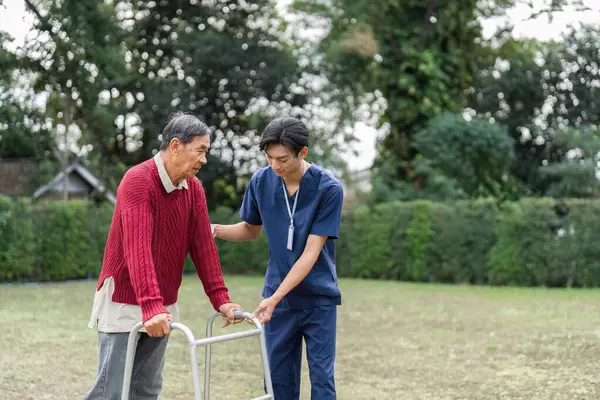 Şefkatli bir sağlık uzmanı yaşlı bir adama yürüteç kullanarak yönünü bulmasında yardımcı olur, sakin yeşil bir ortamda güven ve desteği vurgular..