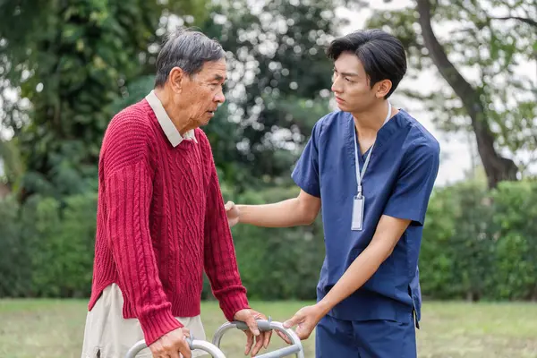 Yaşlı bir adam, bakıcı bir sağlık çalışanı tarafından desteklenir. Bir yürüteçle güzel bir açık alanda gezinirler. Güven ve dostluk gösterirler..
