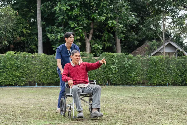 Şefkatli bir sağlık çalışanı, mutlu bir şekilde yaşlı bir adamı tekerlekli sandalyeye itiyor. İkisi de huzurlu bir bahçenin güzel çevresini takdir ediyor..