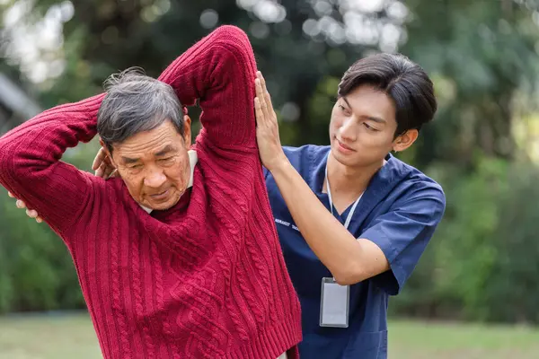 Kendini adamış genç bir bakıcı, yaşlı bir adama yemyeşil bir bahçede esneme egzersizi yapması için destek olur..