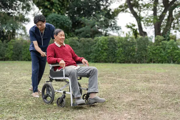 Bir hemşire yardımcısı mutlu bir şekilde tekerlekli sandalyedeki yaşlı bir adamı güzel bir bahçeye itiyor. Doğal ortamdaki hareketlilik ve özen sevincini vurguluyor..